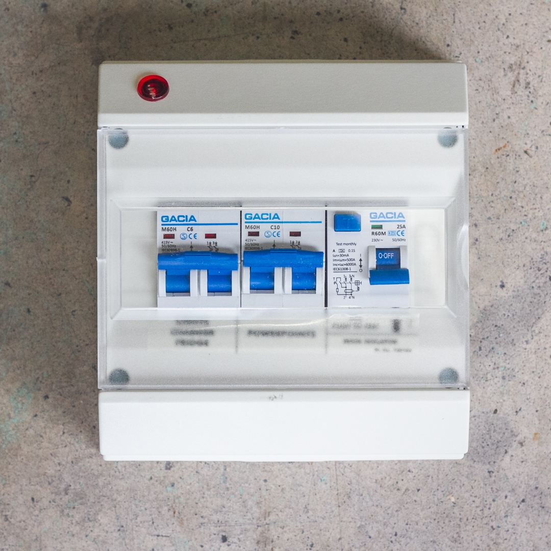 The Consumer Unit - 25A RCD, 10A & 6A double pole MCB, designed for a white campervan, features three blue-capped circuit breakers and a red indicator light, set against a textured wall.