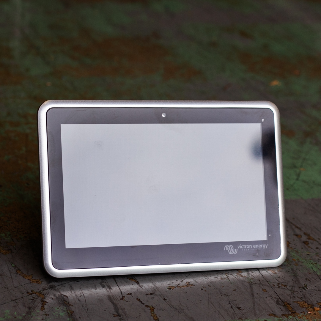 A small touchscreen display device featuring the Victron Energy logo rests on a dark, worn green surface— it's the sleek Victron Ekrano GX Monitoring System.
