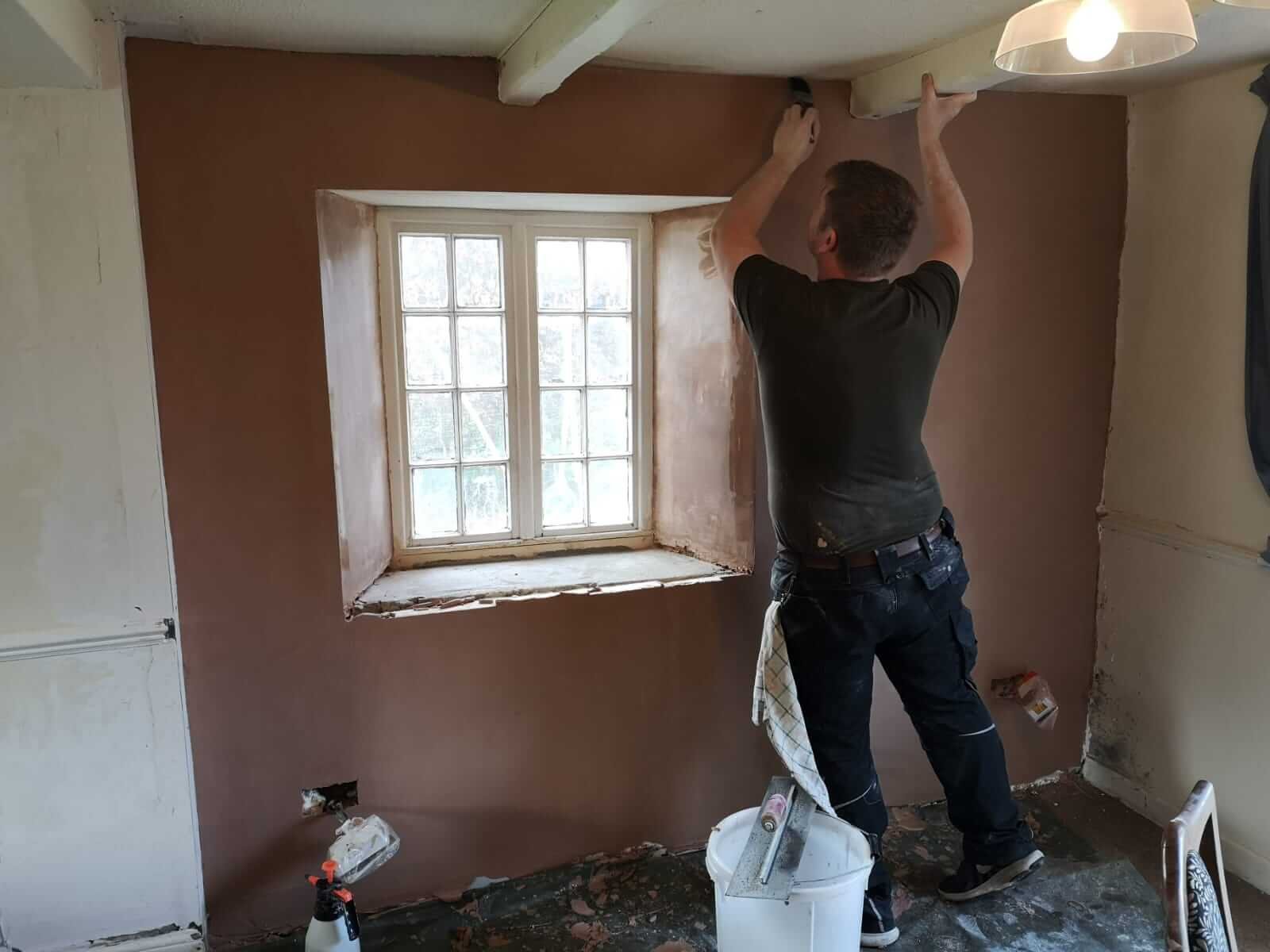 Person smoothing plaster on a wall around a window in a partially renovated room. Construction tools and materials nearby.