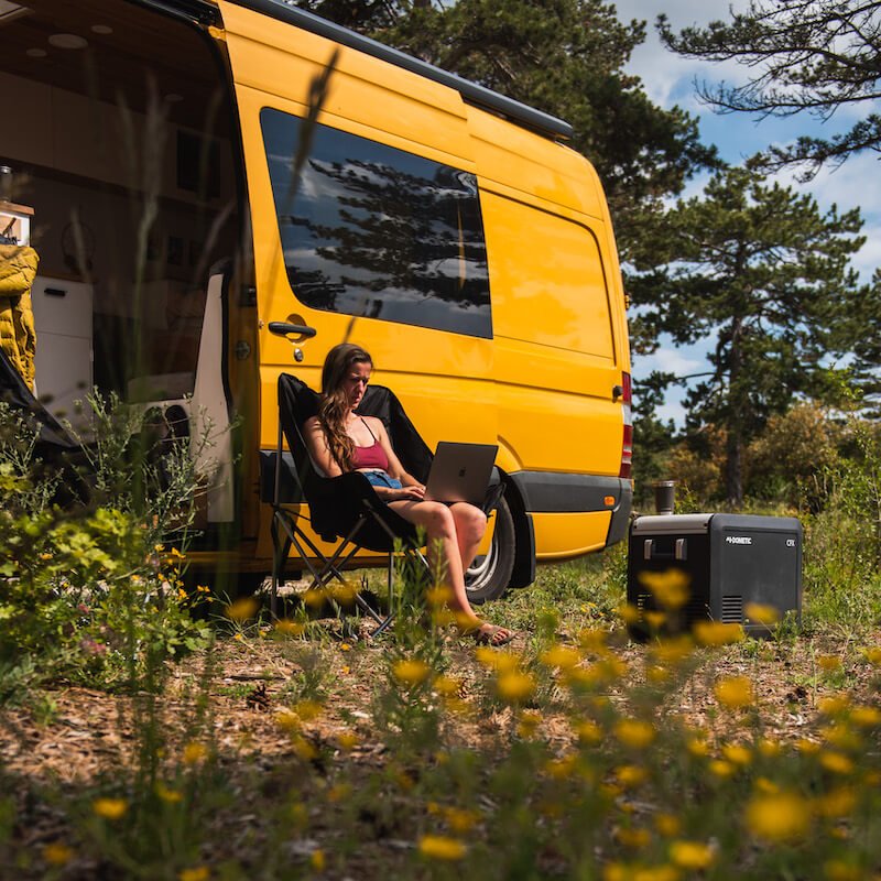 A person works on a laptop in a camping chair next to a yellow van and a portable generator, with trees and flowers nearby, alongside their trusty Dometic CFX3 35 - Compressor Cool Box - 36L Portable Fridge Freezer.