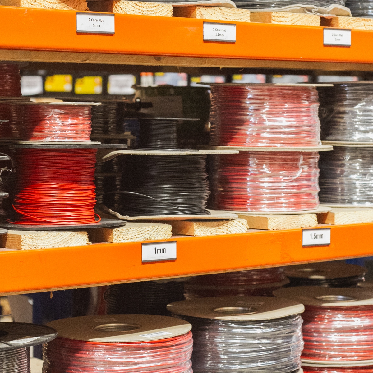 Spools of wire in various colors, including Black DC Power Cable, are neatly arranged on orange shelves in the hardware store, each labeled with different sizes and types.