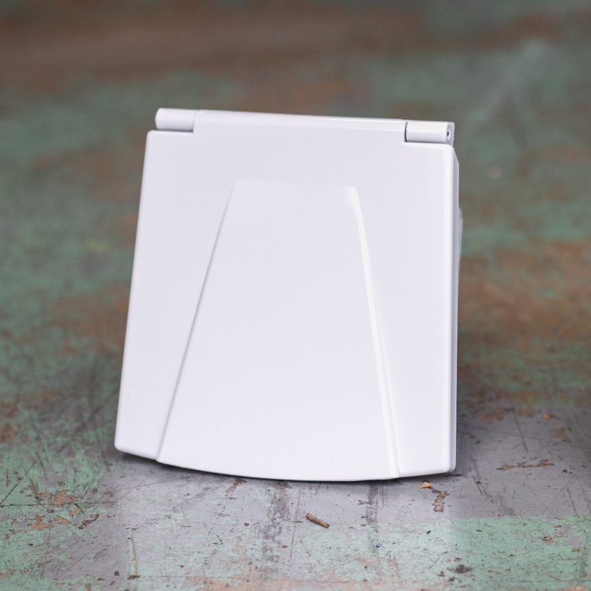 A white, clamshell portable gaming console is closed and positioned facing outward on a weathered surface, with a Mains Hook Up inlet socket - 16A hook-up socket for campervans (white) visible at the side.