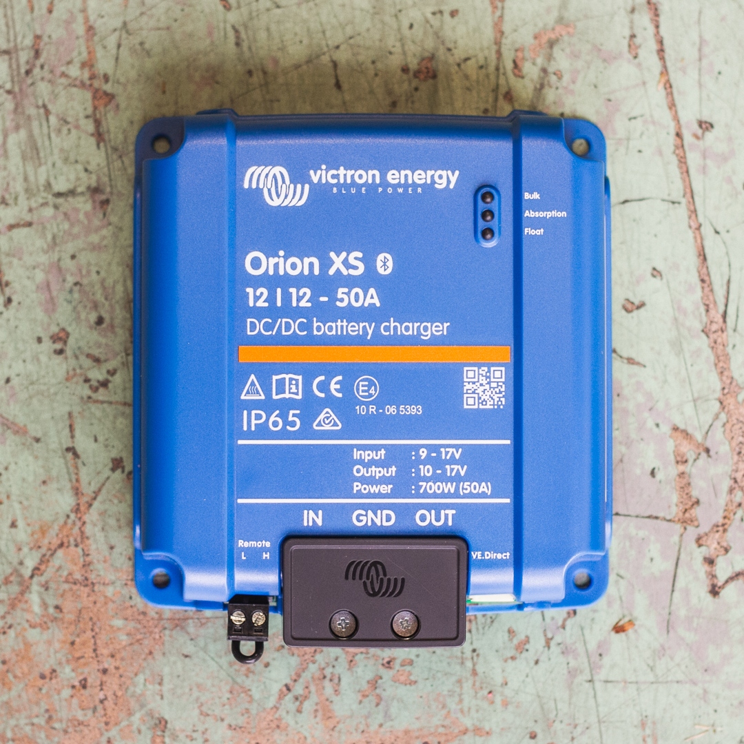 On a weathered surface lies the blue Orion XS 12/12-50A DC-DC Battery Charger - Victron Non-Isolated Smart BuckBoost, complete with its specifications and ports.
