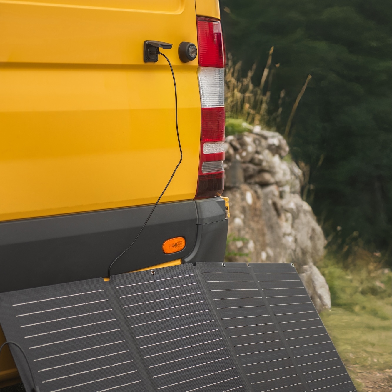 A yellow van is parked outdoors near rocks and greenery, connected to a portable solar panel using the Scanstrut Ventura - XT60 Solar Connection.