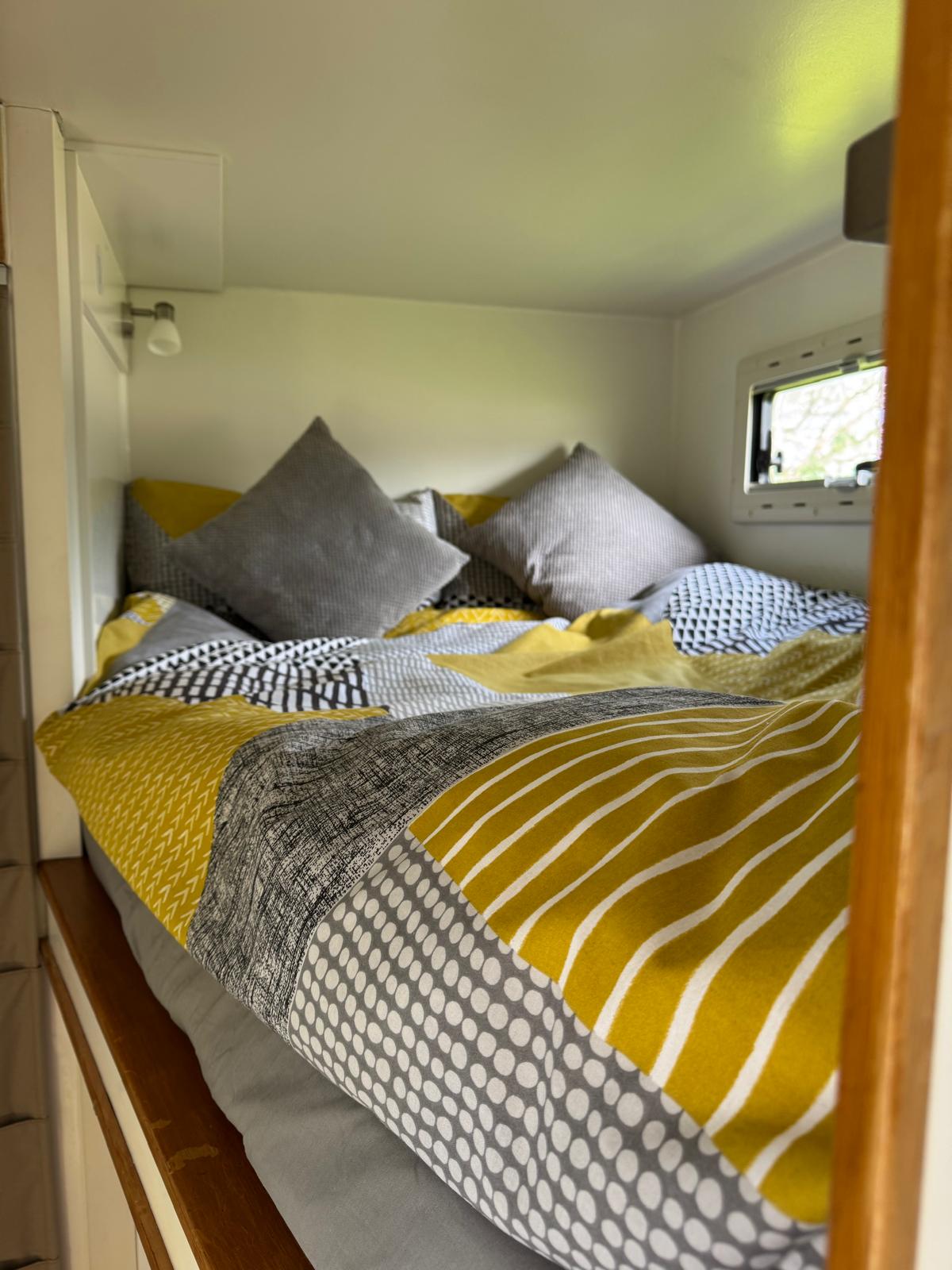 A cozy bed with yellow, gray, and white patterned bedding and pillows sits inside a small, bright room with a window—perfect for relaxing after an adventurous day out in your Iveco Trakker 4x4.