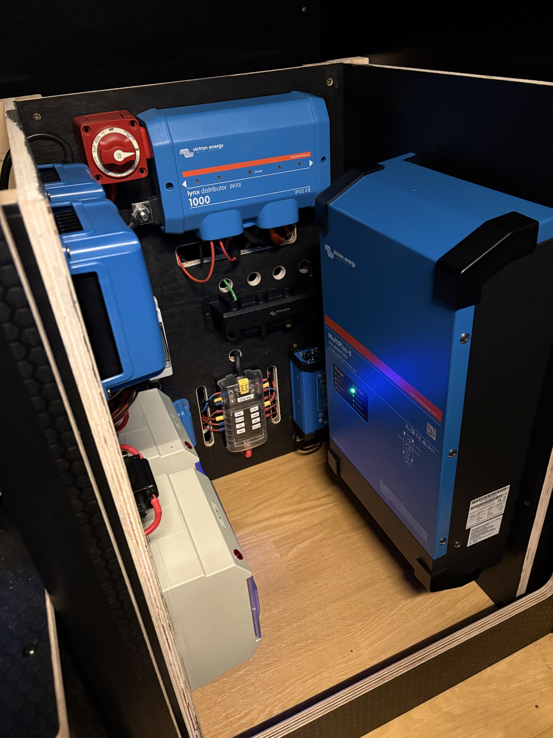 Blue electrical equipment and battery system by Sam Kidd neatly installed in a wooden enclosure with organised wiring and a fuse panel.