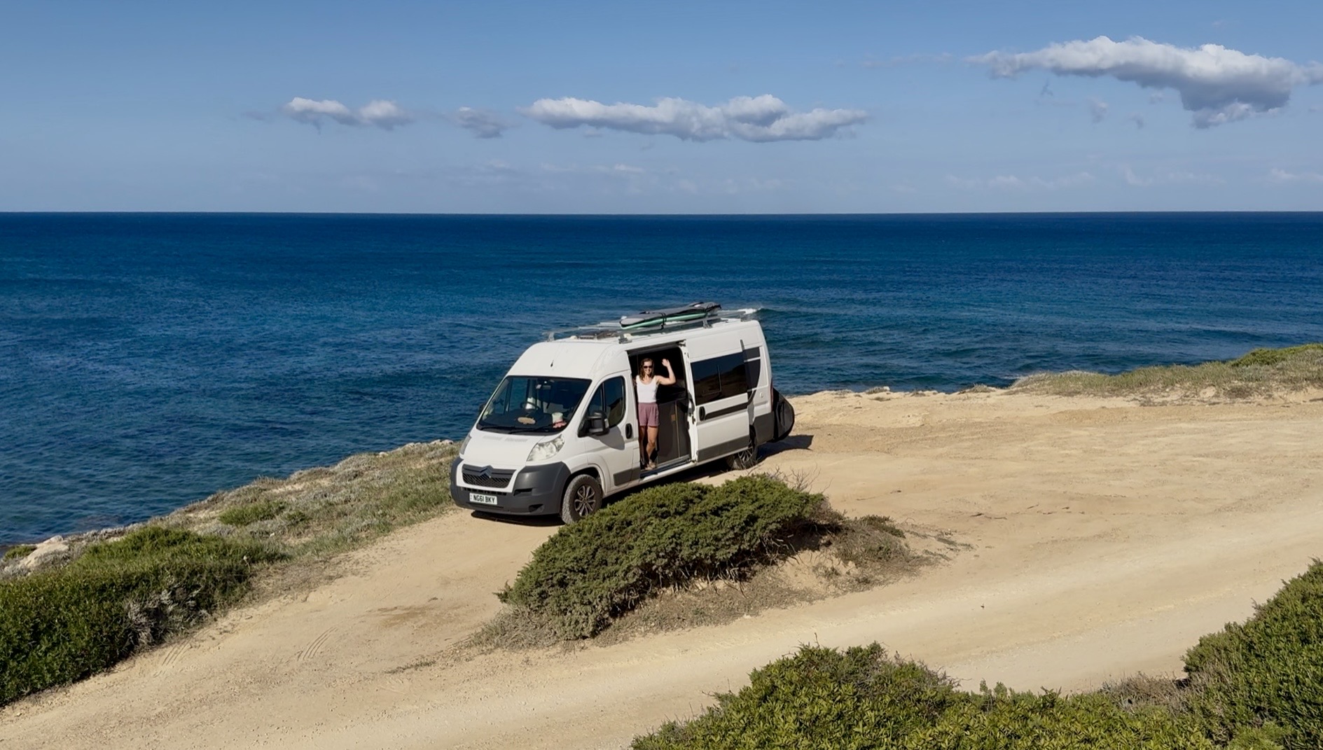 LJ waving from the sliding door of their self converted van which is parked on a cliff overlooking the sea