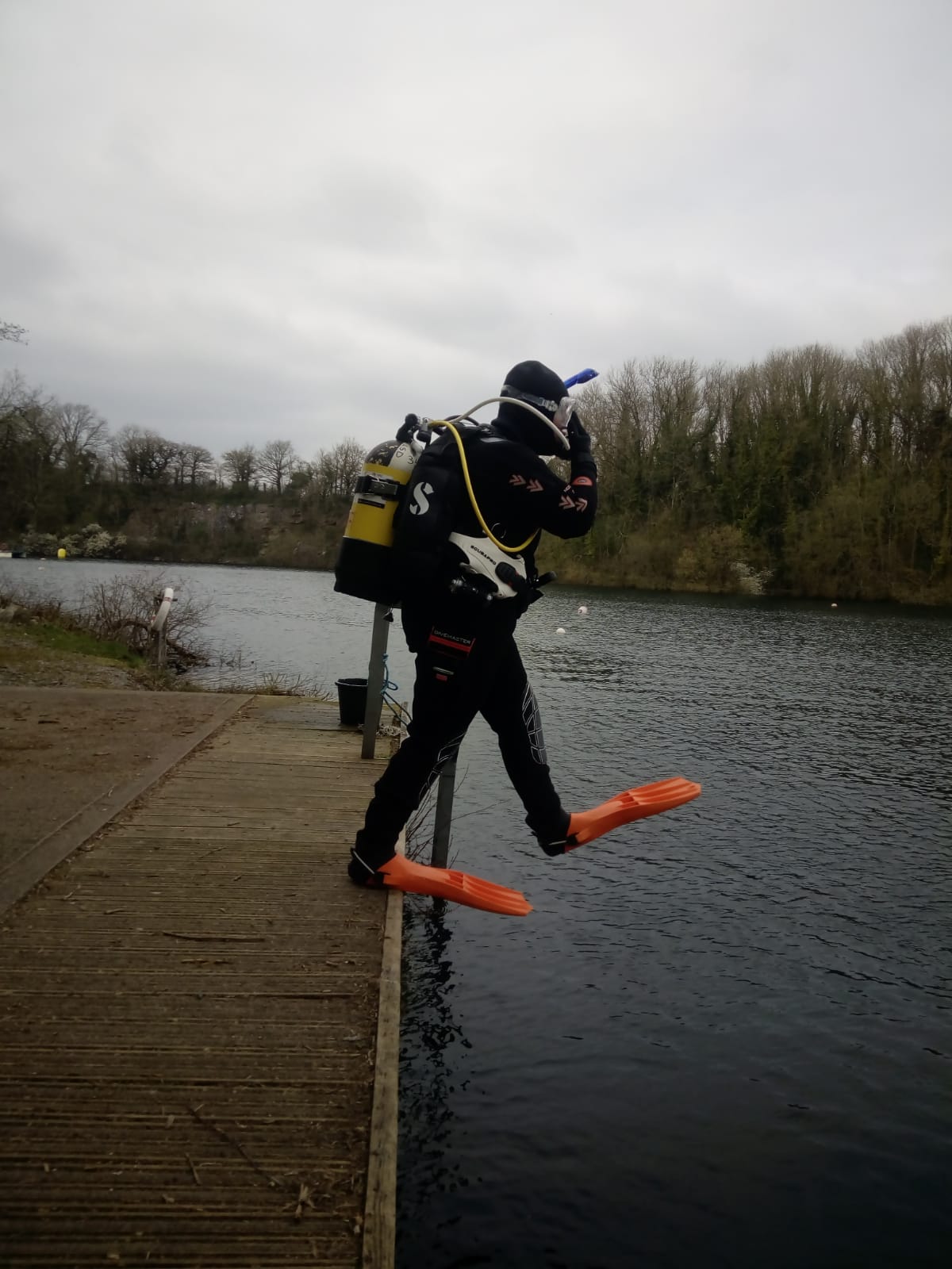 Martina in full cold water scuba gear with orange fins doing a deep entry at a diving quarry in Somerset UK