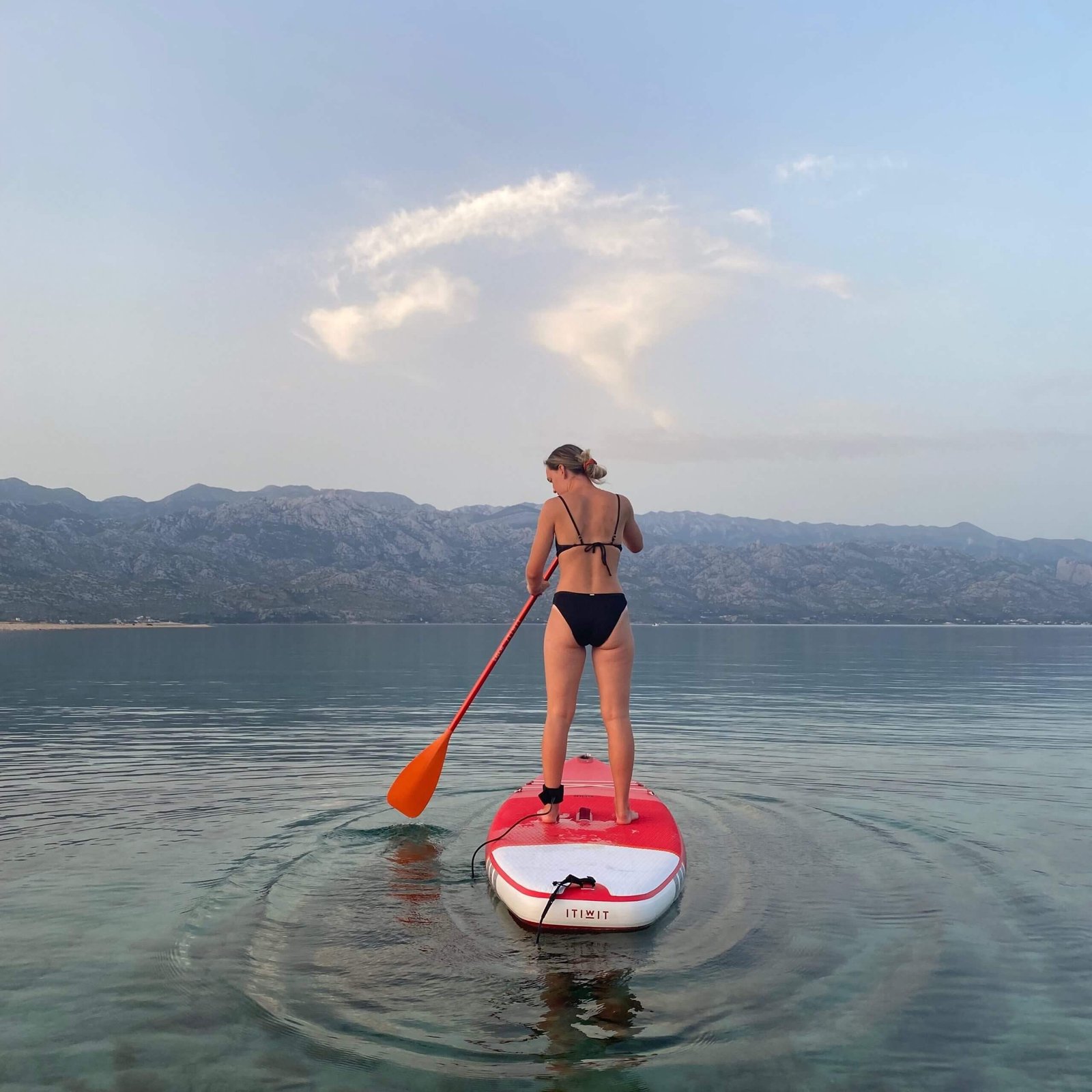 Becky standing on a white and red sup wearing a black bikini on a lake with mountains in the background