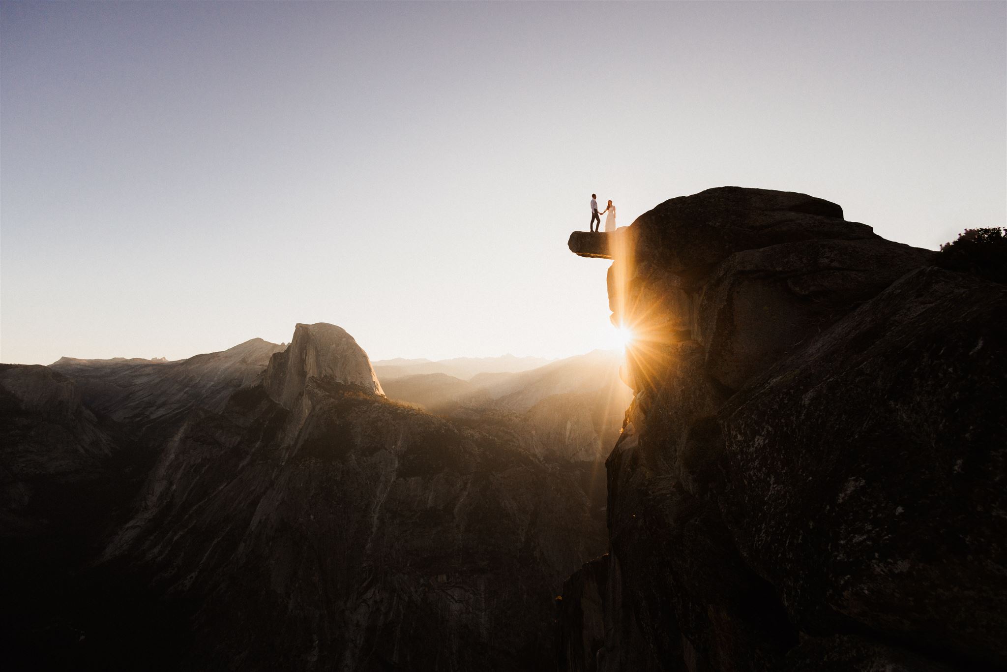 a woman and a man standing on top of a cliff overlooking the sunset