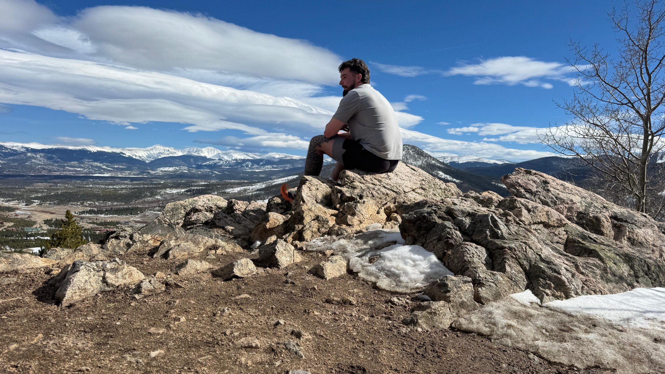 Huey sitting on a rock on top of a mountain surrounded by little snow piles with snow capped mountain peaks in the background